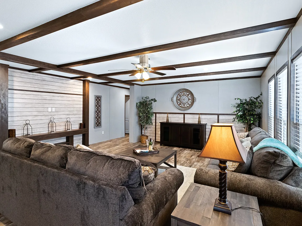 Spacious living room with brown sofas, wooden beams, and a ceiling fan. Soft lighting from a table lamp, decorated with plants and wall art. Cozy and inviting ambiance.