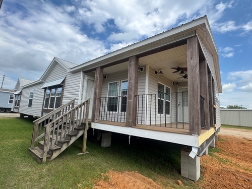 A single-story mobile home with a wooden porch on pillars, steps leading up, under a partly cloudy sky. Surroundings are grassy with patches of bare earth.