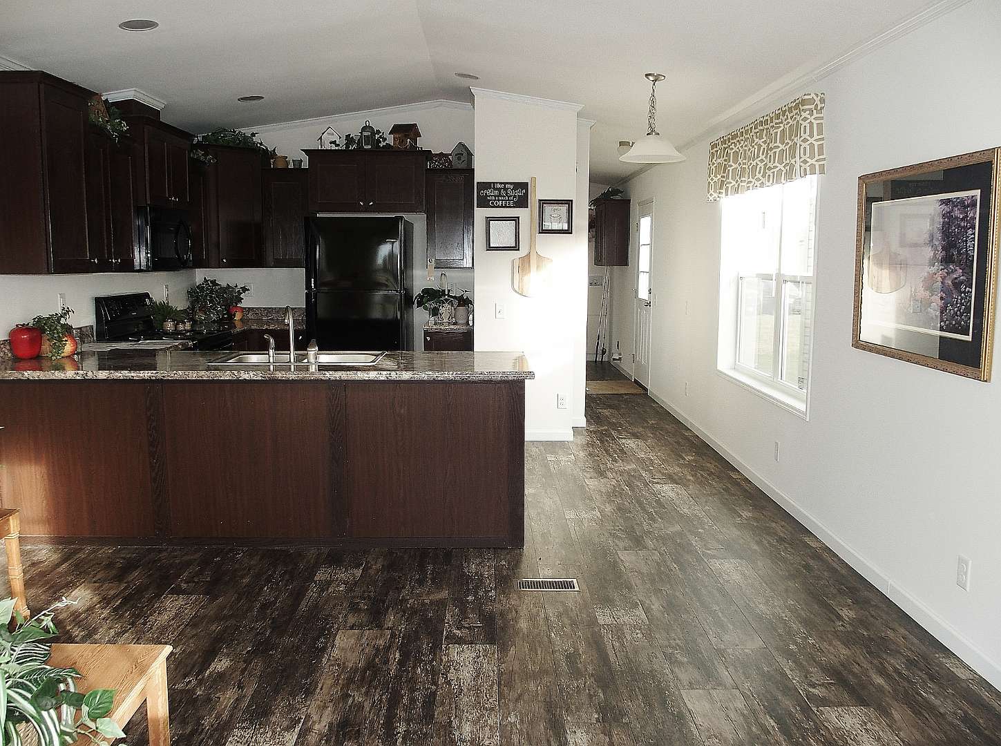 Spacious kitchen with dark wood cabinets, marble countertop, and stainless steel appliances. Sunlit wood floor leads to a bright windowed hallway. Cozy ambiance.