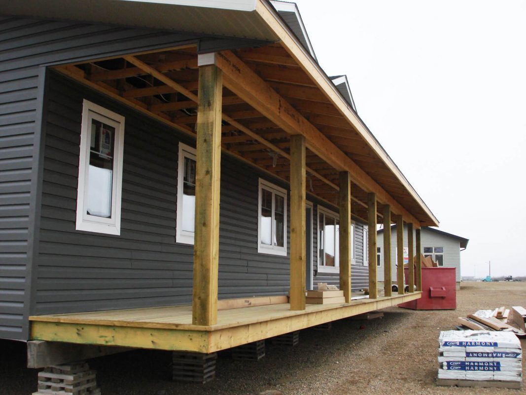 Elevated wooden porch under construction on a gray-sided house. Exposed beams and unfinished posts are visible. Materials are stacked nearby.