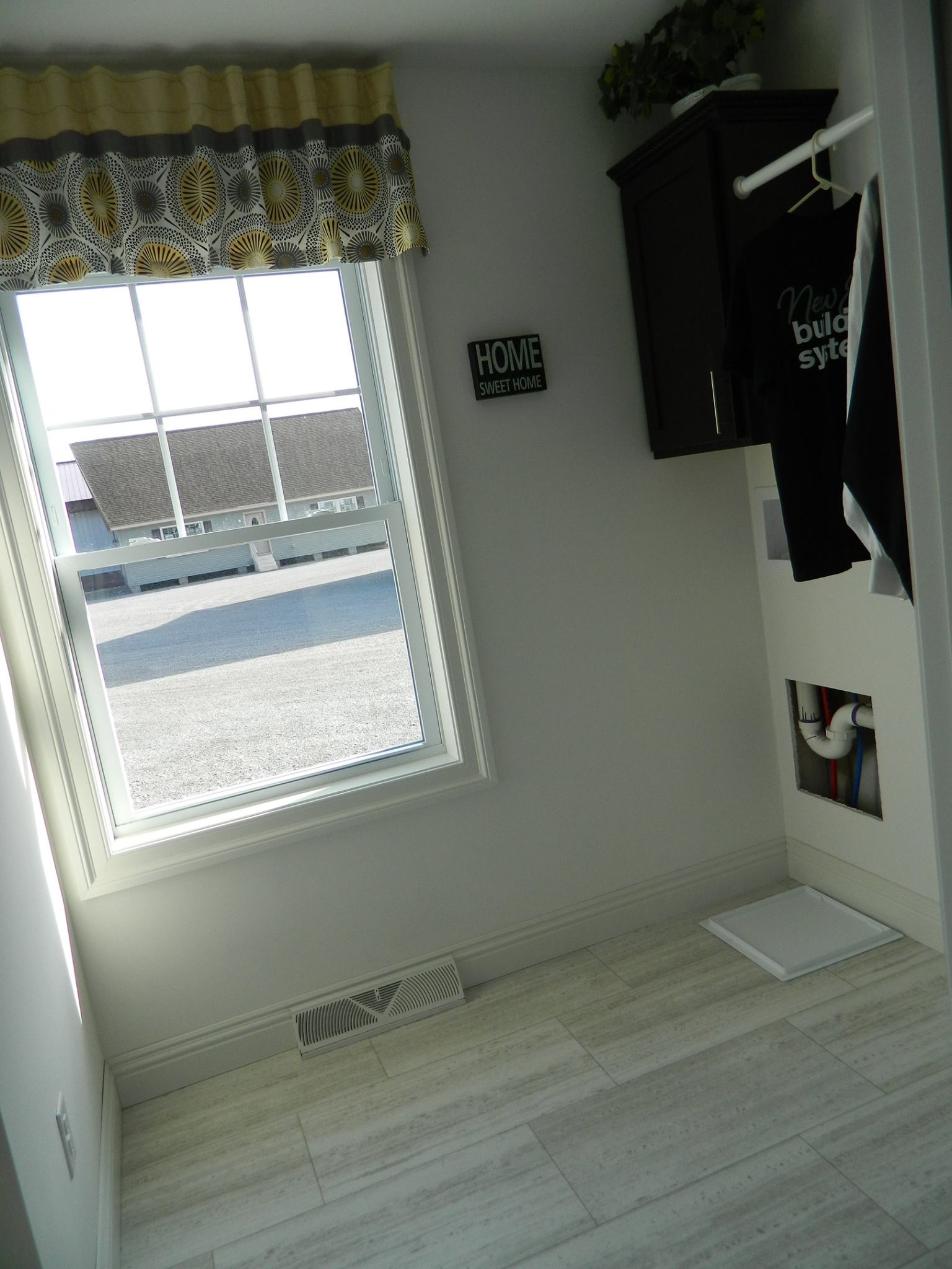 A bright laundry room with a window featuring patterned curtains, a "Home Sweet Home" sign, dark cabinets, and a white laundry basket on tiled floors.