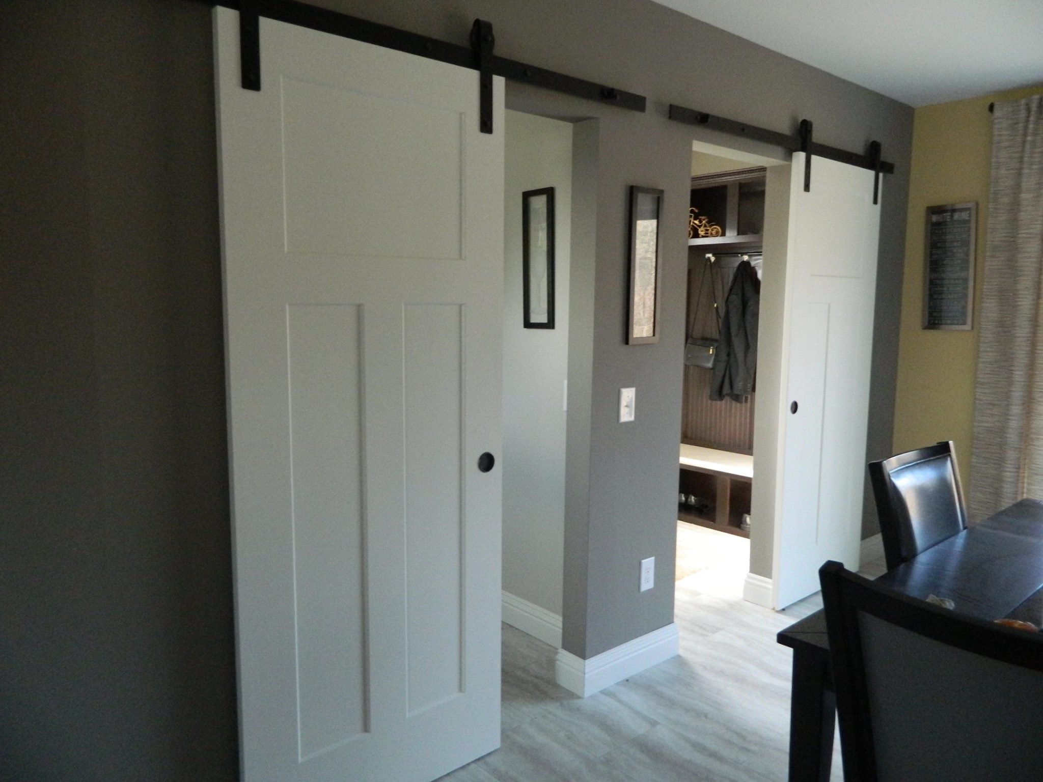 Modern room with white sliding barn doors on black metal tracks. Doors open to reveal a closet with coats. Gray and beige walls add a sleek tone.