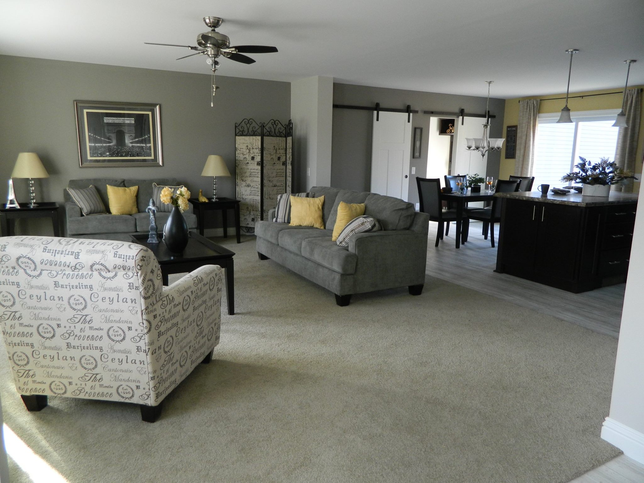 Modern living room with gray sofa, yellow cushions, and an armchair with text print. A dining area and kitchen with pendant lights are visible. Cozy ambiance.