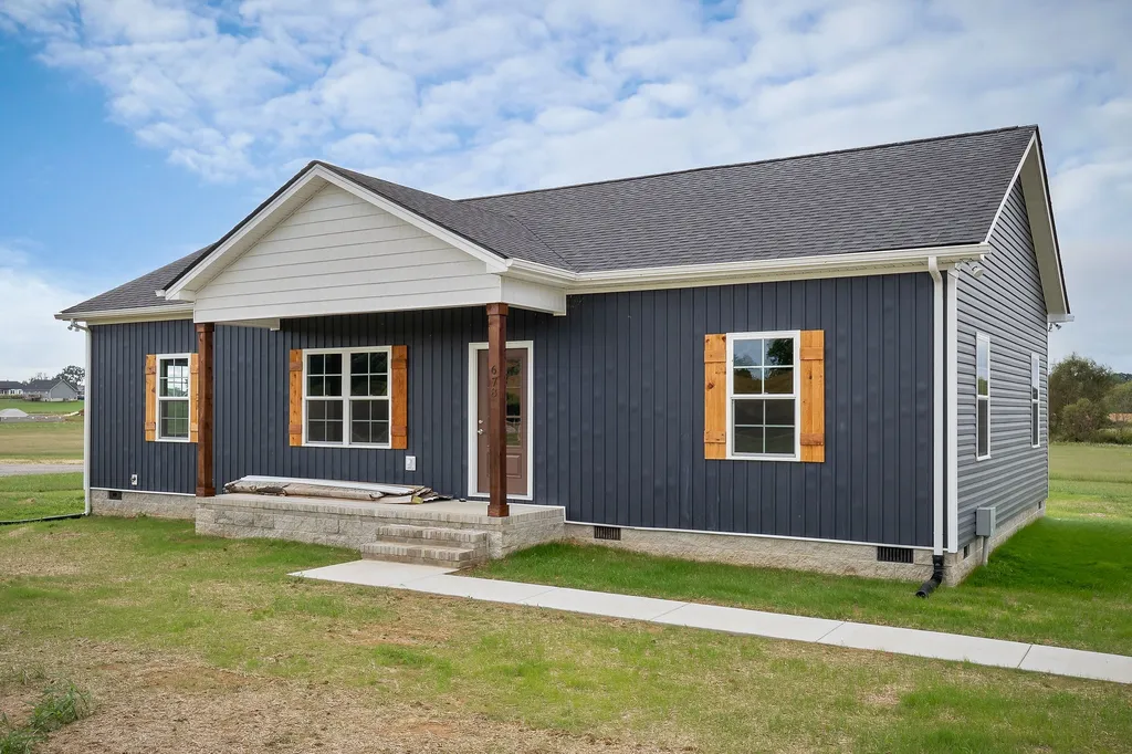 A modern, single-story house with dark blue siding, a gray roof, and wooden shutters. The entrance features two wooden columns. The setting is a grassy area.