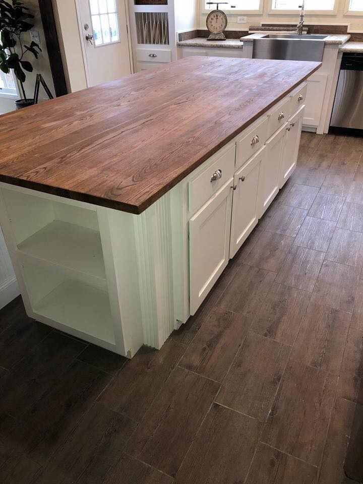 A kitchen island with a wooden countertop and white cabinets is centered on a dark wooden floor. Nearby, there's a stainless steel sink and large windows.