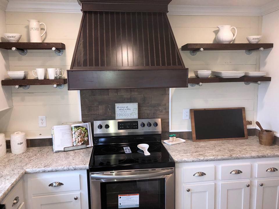 A cozy kitchen features a stainless steel stove with a dark wooden range hood. Open shelves display white dishes, cups, and pitchers. A small chalkboard and a wooden utensil holder sit on the countertop, conveying a rustic and homey feel.