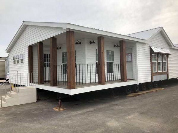 A mobile home with white siding and a metal roof features a raised porch supported by wooden pillars. Black railings and outdoor lights add charm.