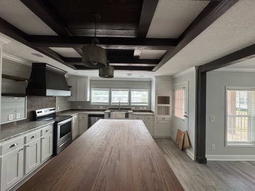 Spacious modern kitchen with a long wooden island, white cabinets, stainless steel appliances, large windows, exposed beams, and calm neutral tones.