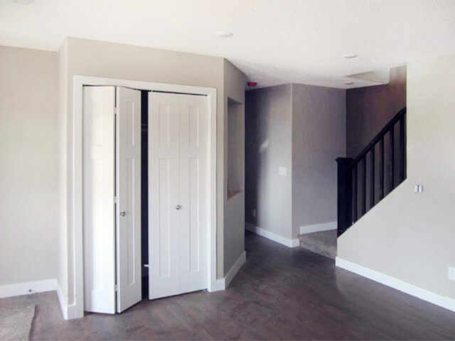 A modern, empty living room with gray walls and dark wood flooring. A partially open white closet door is on the left, next to dark wooden stairs.