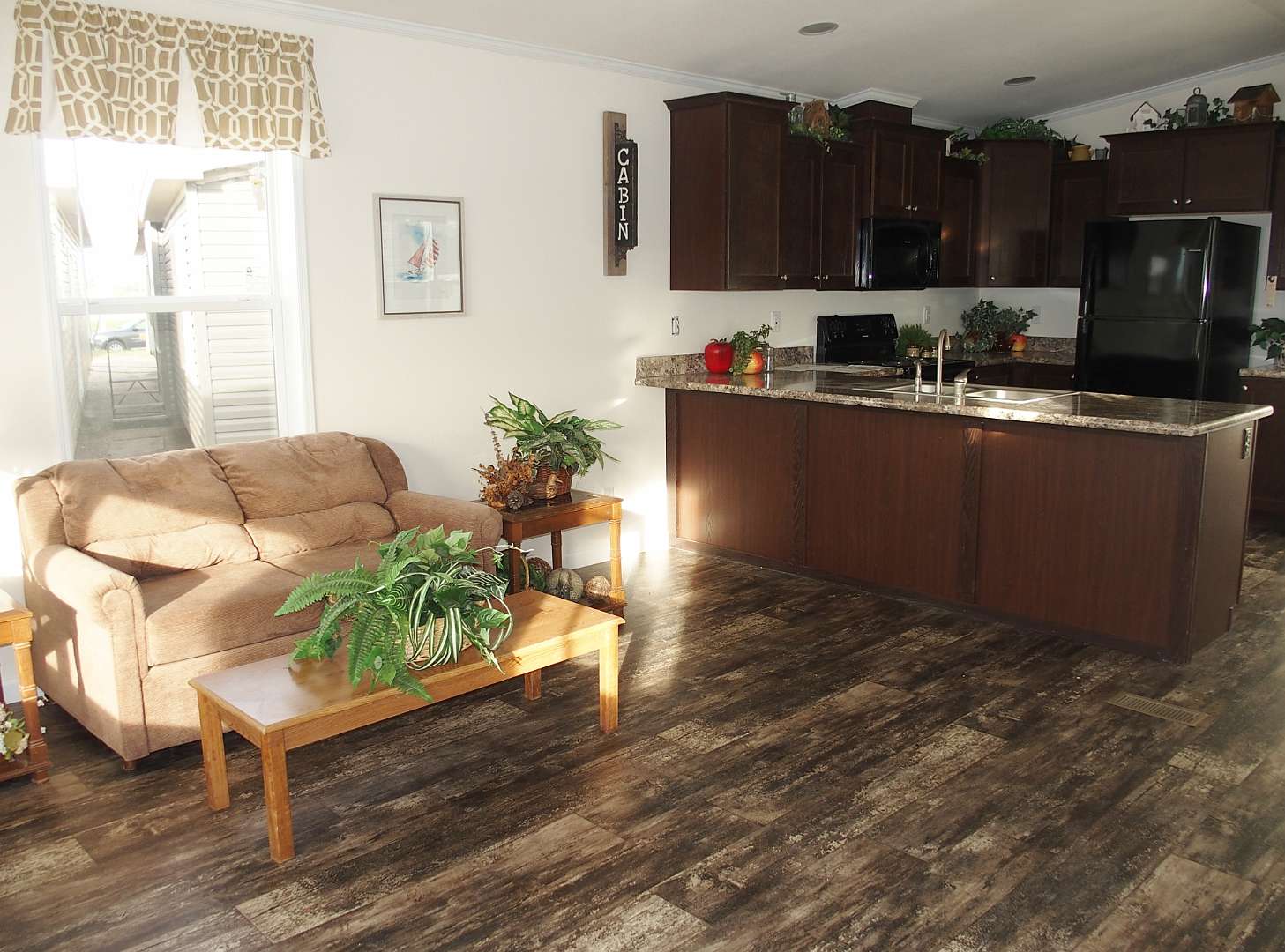 Warm, cozy living room with a tan sofa, wooden table, and lush plants. A modern kitchen with dark cabinets and marble countertops is visible in the background.