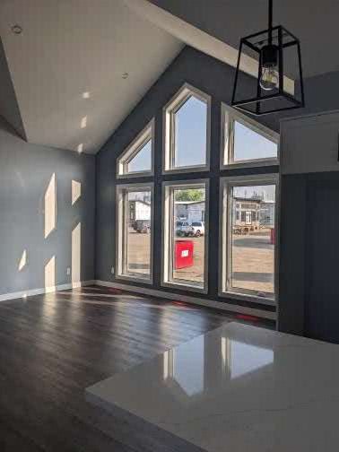 Sunlit modern room with large triangular windows, casting geometric shadows on gray walls. A black pendant light hangs from a high ceiling.