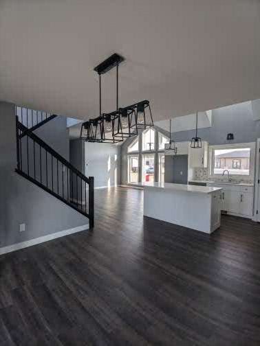 Modern, spacious interior with dark wood floors, a black metal staircase, and a large chandelier. Bright kitchen features white cabinets and an island.