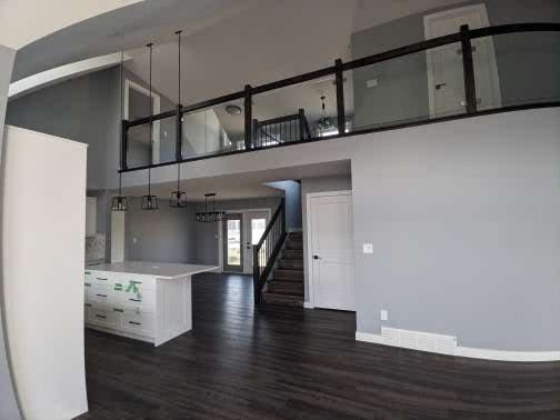 Modern interior with a high ceiling, dark wood flooring, spacious kitchen island, and pendant lights. Glass railings overlook the minimalist design, creating an open, airy feel.
