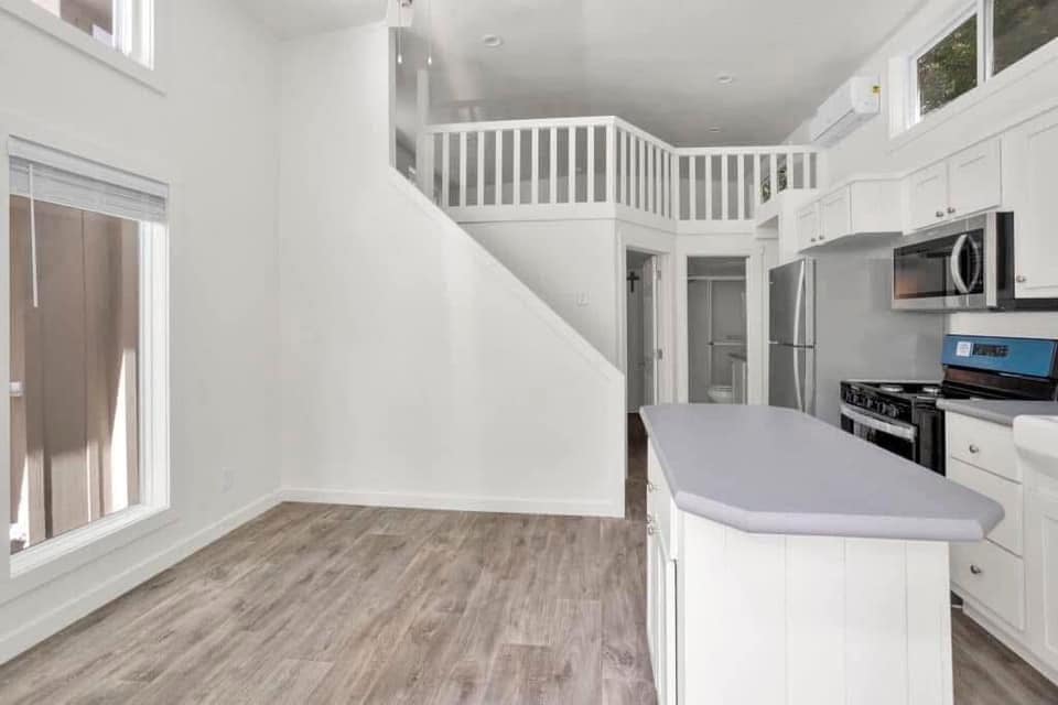 Spacious modern kitchen with white cabinets, gray countertops, and stainless steel appliances. Loft overhead, wood floors, large windows on the left. Airy and bright.
