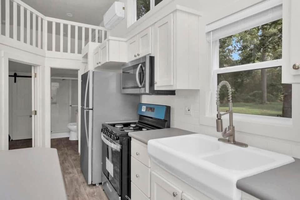 Bright kitchen in a tiny house, featuring white cabinets, a farmhouse sink, stainless steel appliances, and a window view of greenery. Cozy, modern feel.