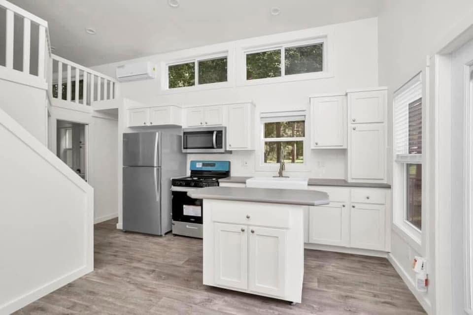 Compact, modern kitchen with white cabinets and stainless-steel appliances, featuring a small island. Large windows allow ample natural light.