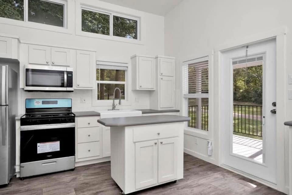 Small, bright kitchen with white cabinets, stainless steel appliances, and a central island. Large windows offer views of lush greenery. Modern and airy.