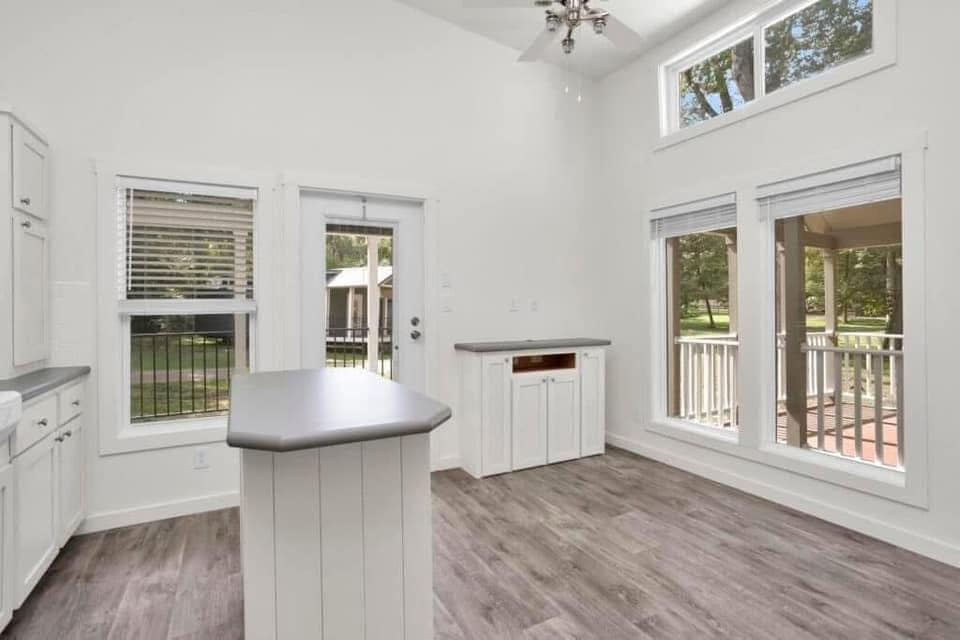 Bright kitchen with white cabinets, a central island, and light wood flooring. Large windows and a glass door lead to a porch with greenery outside.