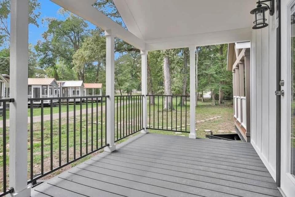 Covered porch with black railing overlooks grassy area with trees. Tiny houses line a distant path. Bright, peaceful atmosphere.