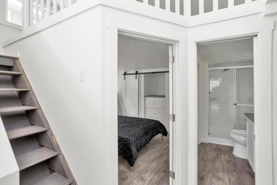 A modern, compact living space features wooden stairs, a small bedroom with a black bedspread, and an adjacent bathroom with a glass shower. The atmosphere is bright and minimalistic.