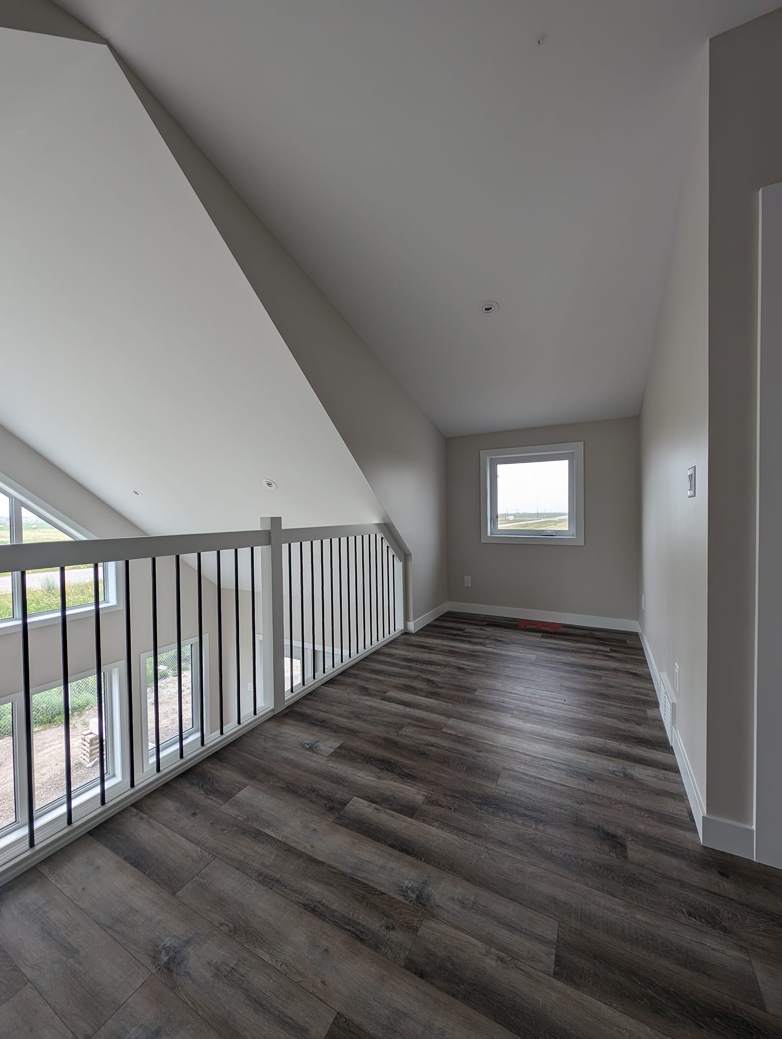 Modern loft space with dark wood flooring, sloped ceiling, and a small window. Metal and wood railing overlooks the floor below, offering an airy feel.
