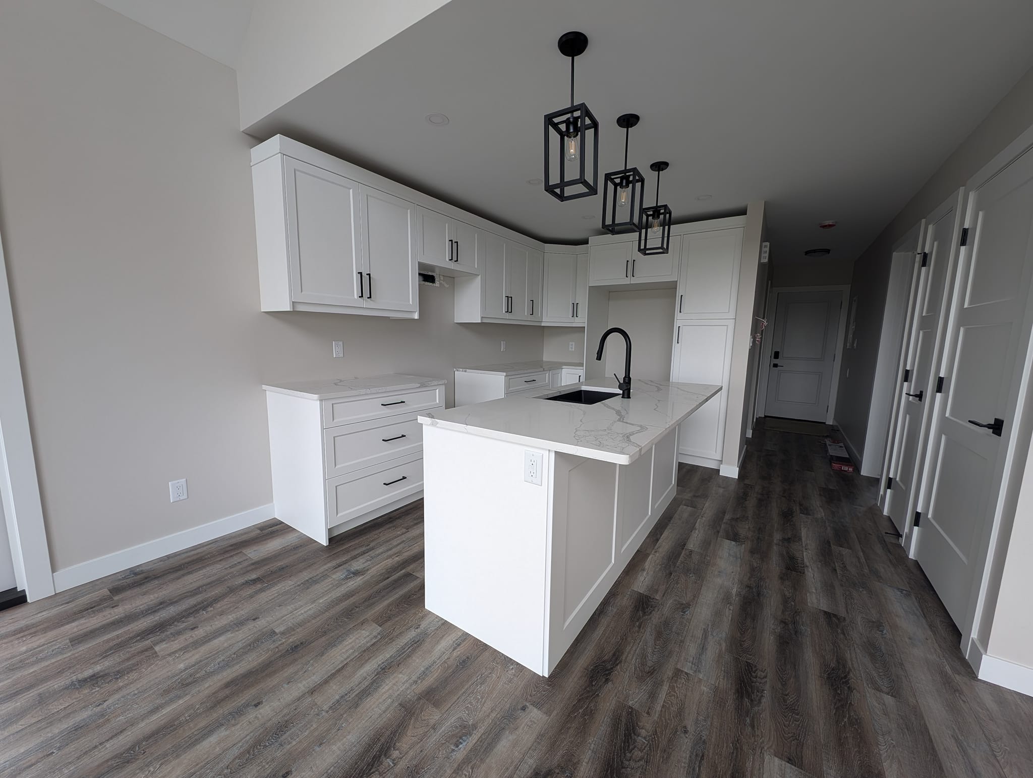 Modern kitchen with white cabinets and a marble island, featuring black fixtures and pendant lights. The wood flooring adds warmth and contrast.