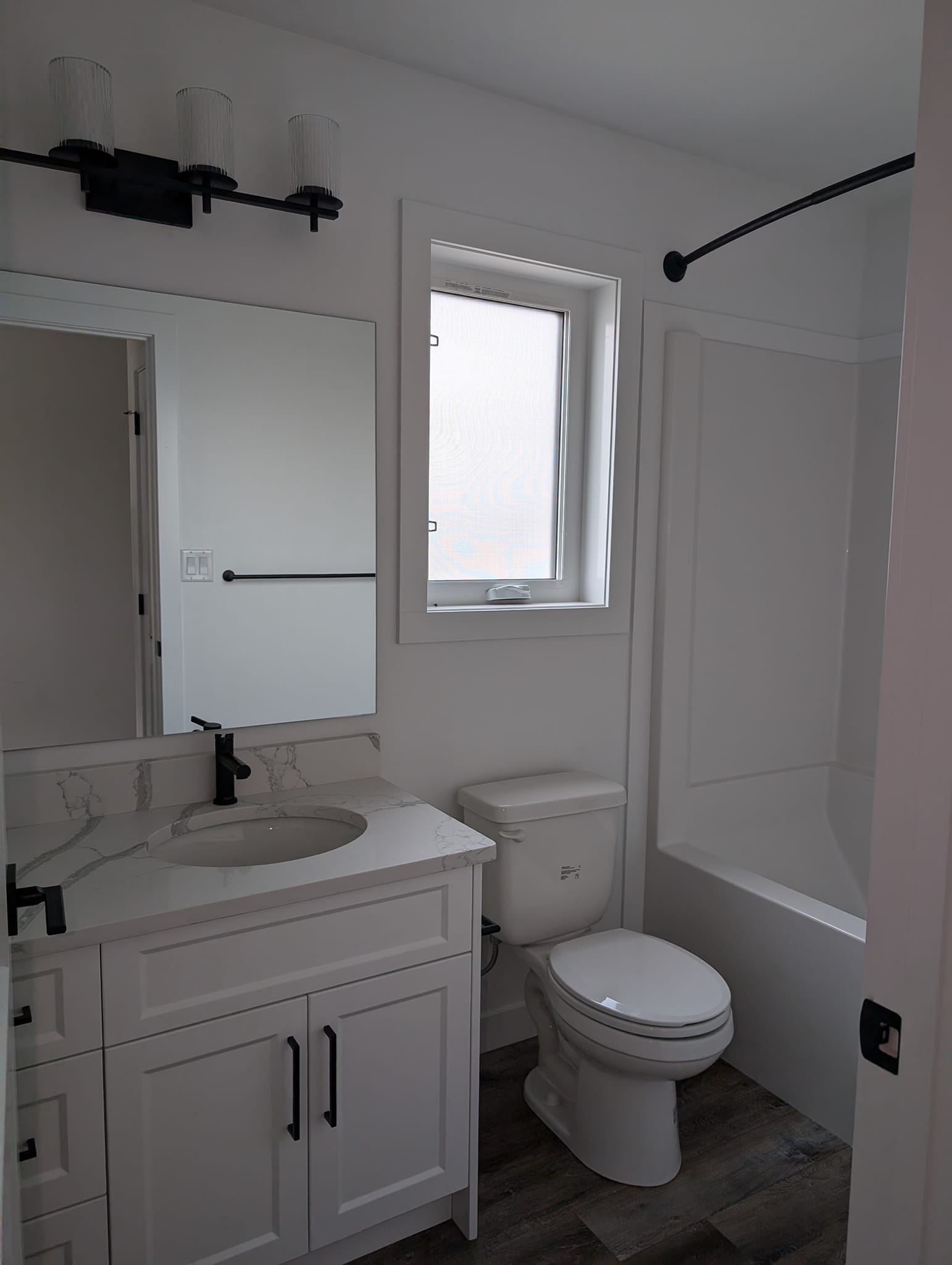 Compact modern bathroom with white cabinetry, black fixtures, marble countertop, mounted mirror, window, and bathtub. Calm, minimalist design.