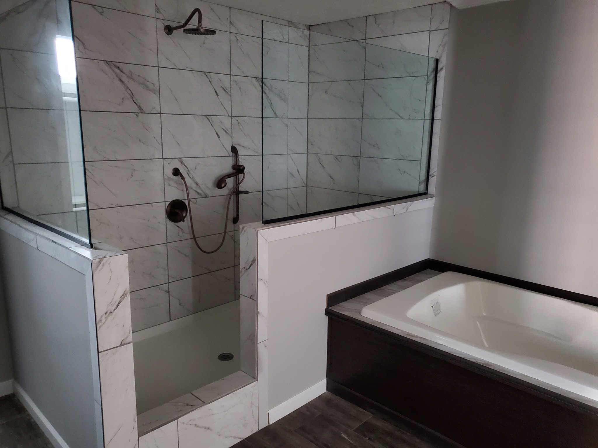 Modern bathroom with a marble-tiled shower featuring glass panels and bronze fixtures. Adjacent is a white bathtub with a sleek dark wooden trim.