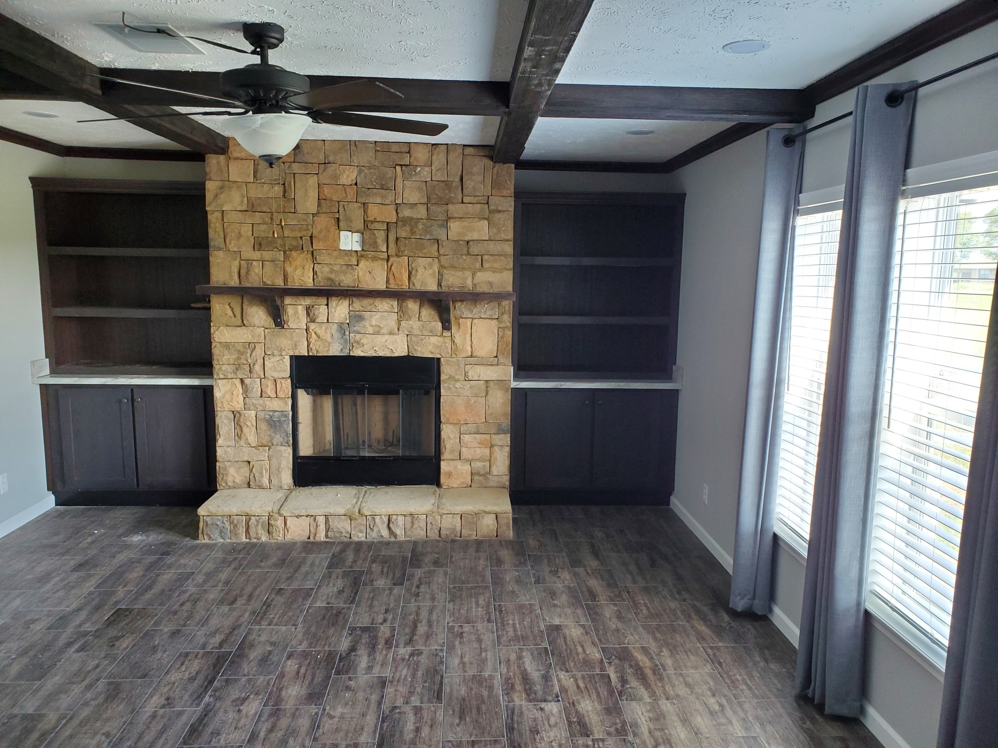 Living room with a stone fireplace, dark wooden cabinets, and beamed ceiling. Two large windows with gray curtains let in natural light. Cozy and rustic.