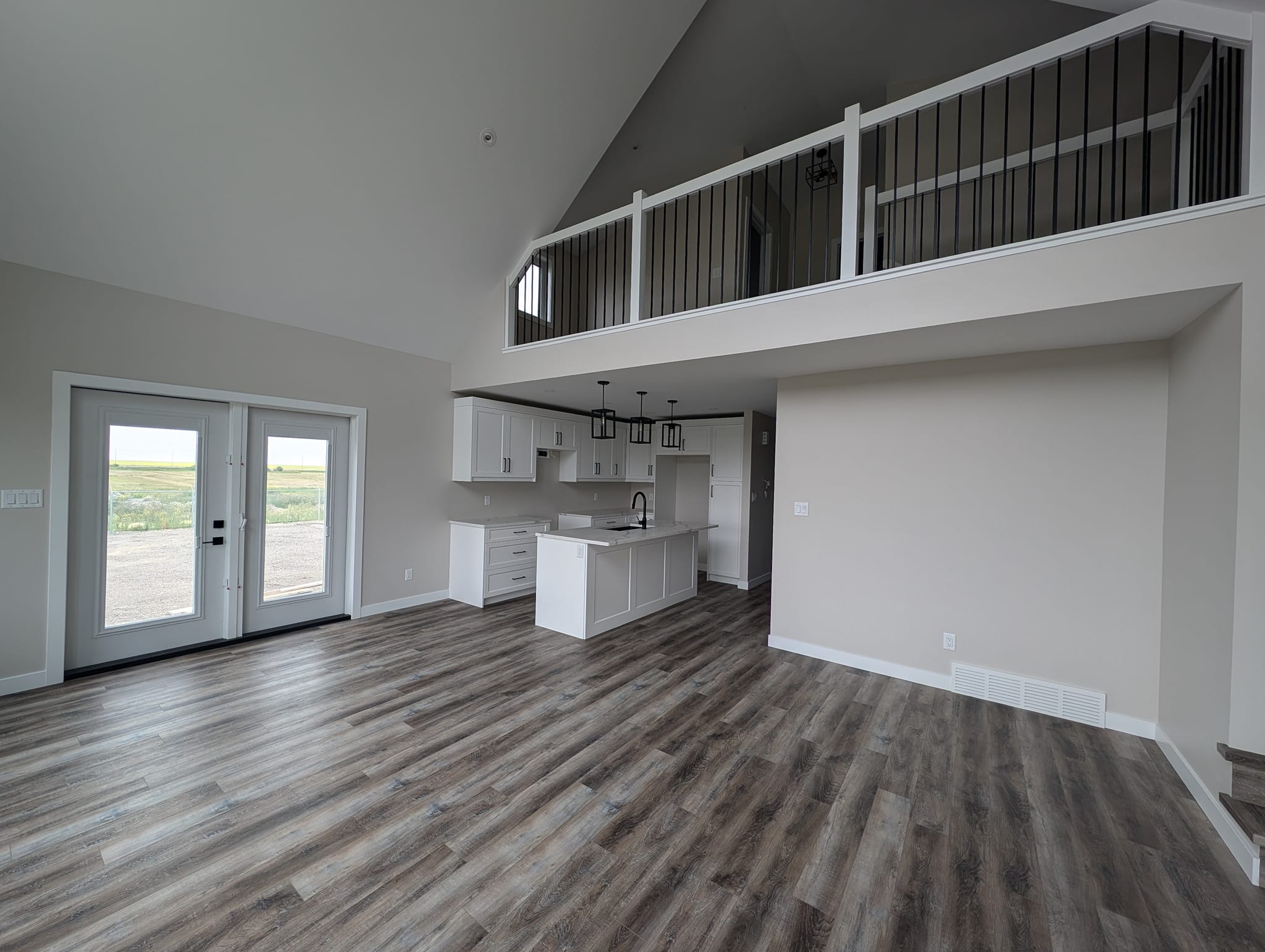 Spacious open-concept living area with high ceilings, wood flooring, and a modern white kitchen. French doors lead to an outdoor view. Airy and inviting.