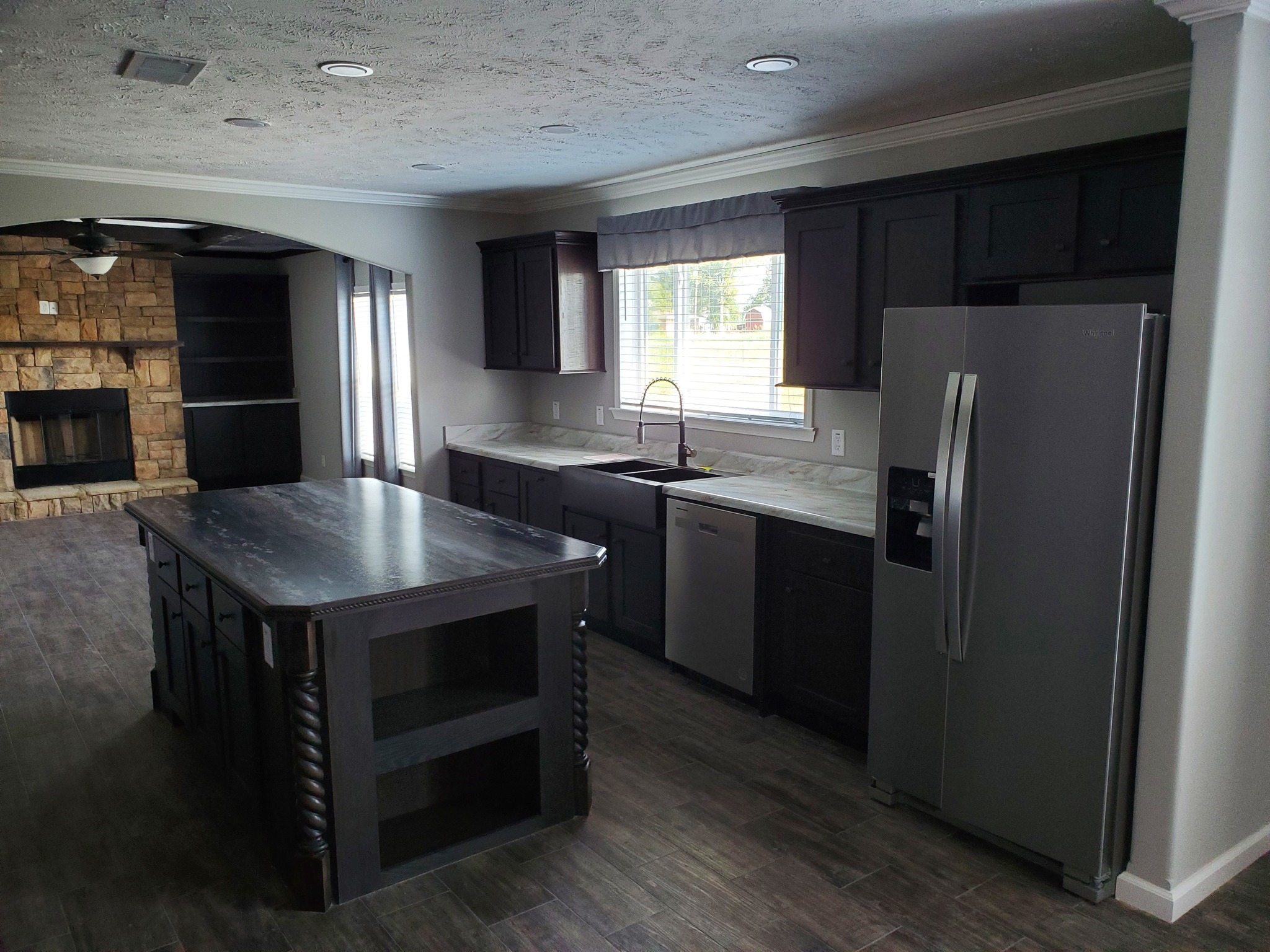 Modern kitchen with dark cabinets, large island, and stainless steel appliances. Open layout connects to a living area with a stone fireplace. Cozy and elegant atmosphere.
