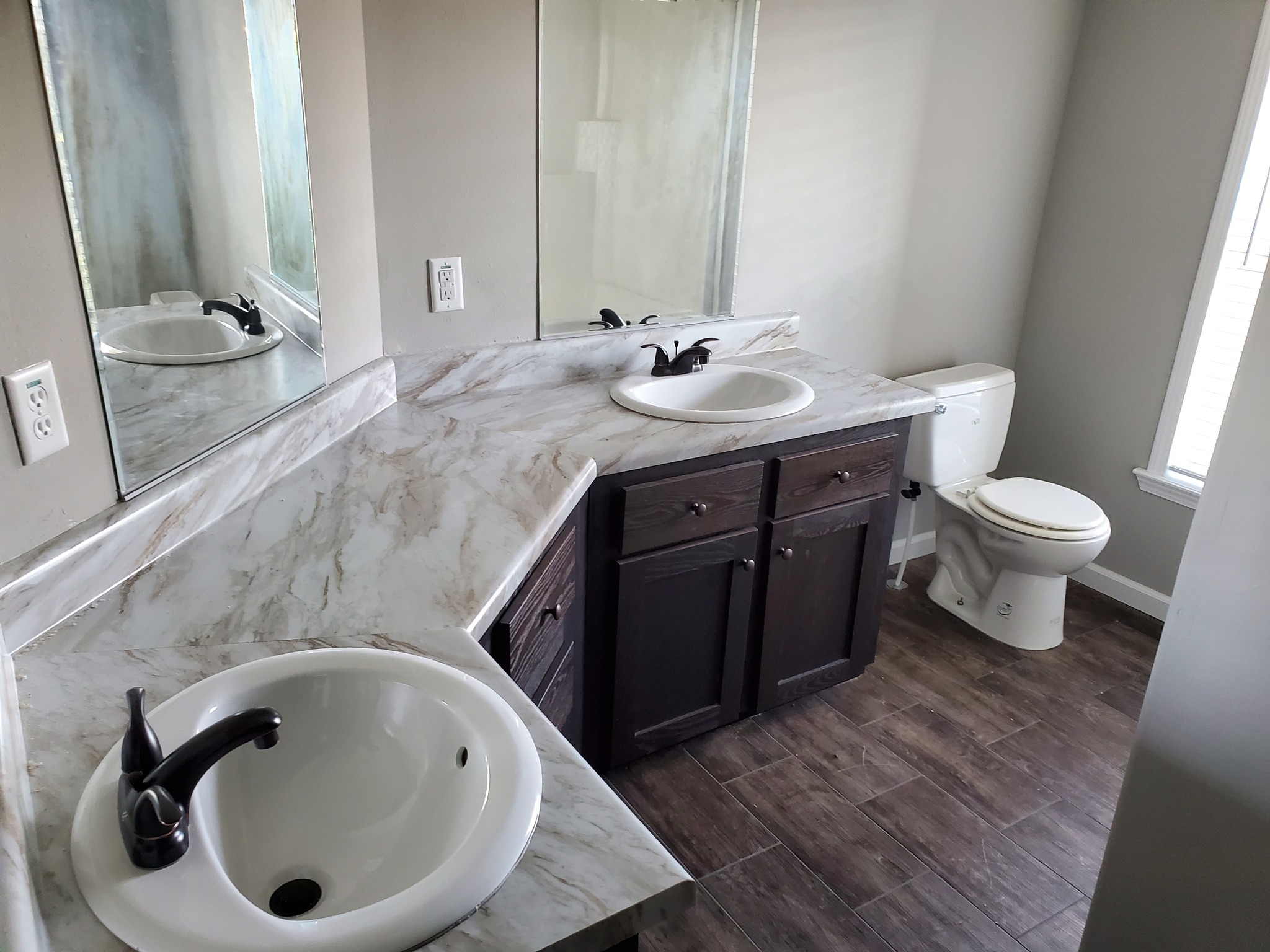 Modern bathroom with marble countertop, dual sinks, and dark wood cabinets. Large mirror above counter, light gray walls, and wood-like tile floor.