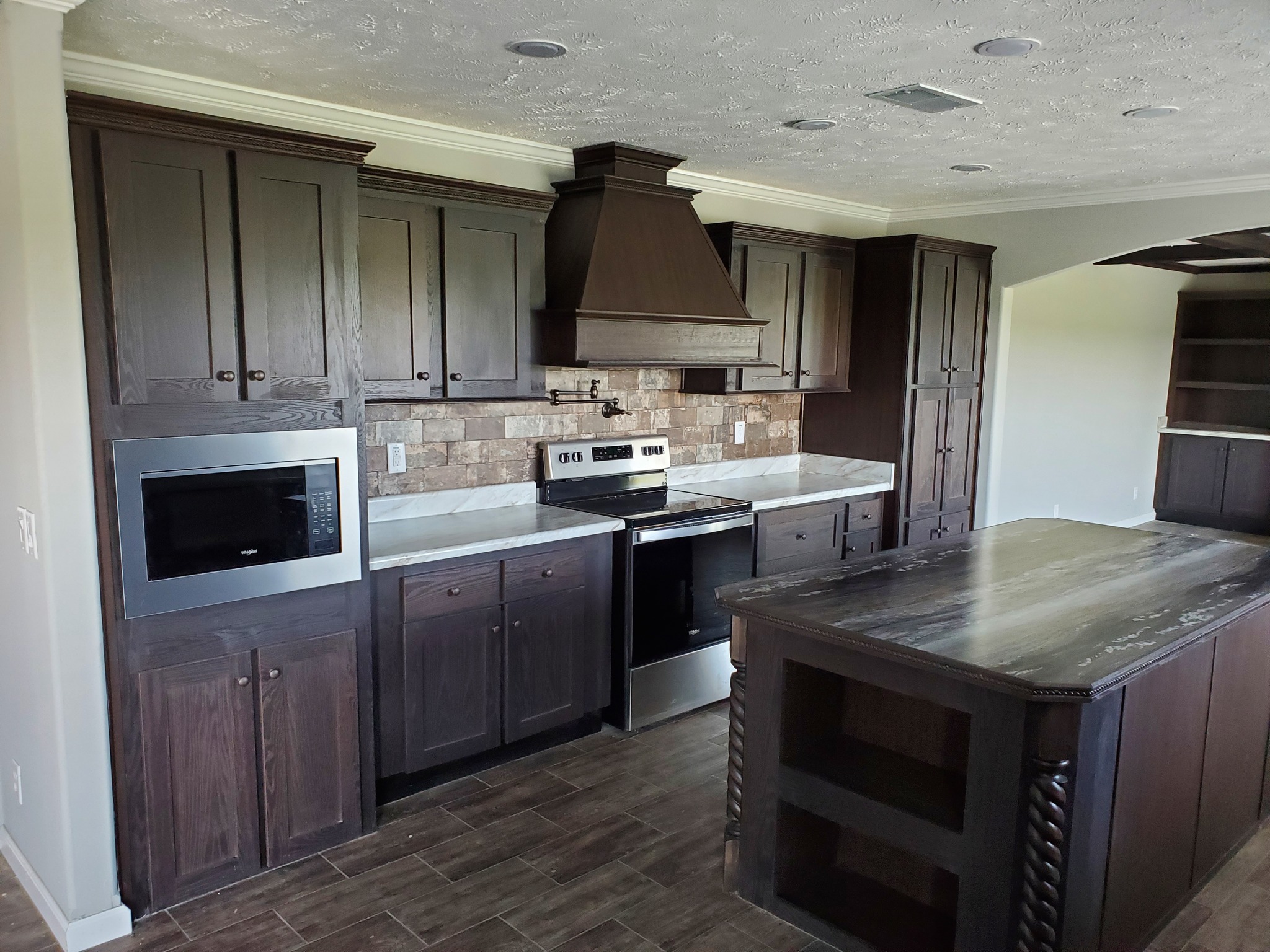 Spacious kitchen with dark wood cabinets, brick backsplash, and stainless steel appliances. Large island with textured countertop. Cozy and modern.