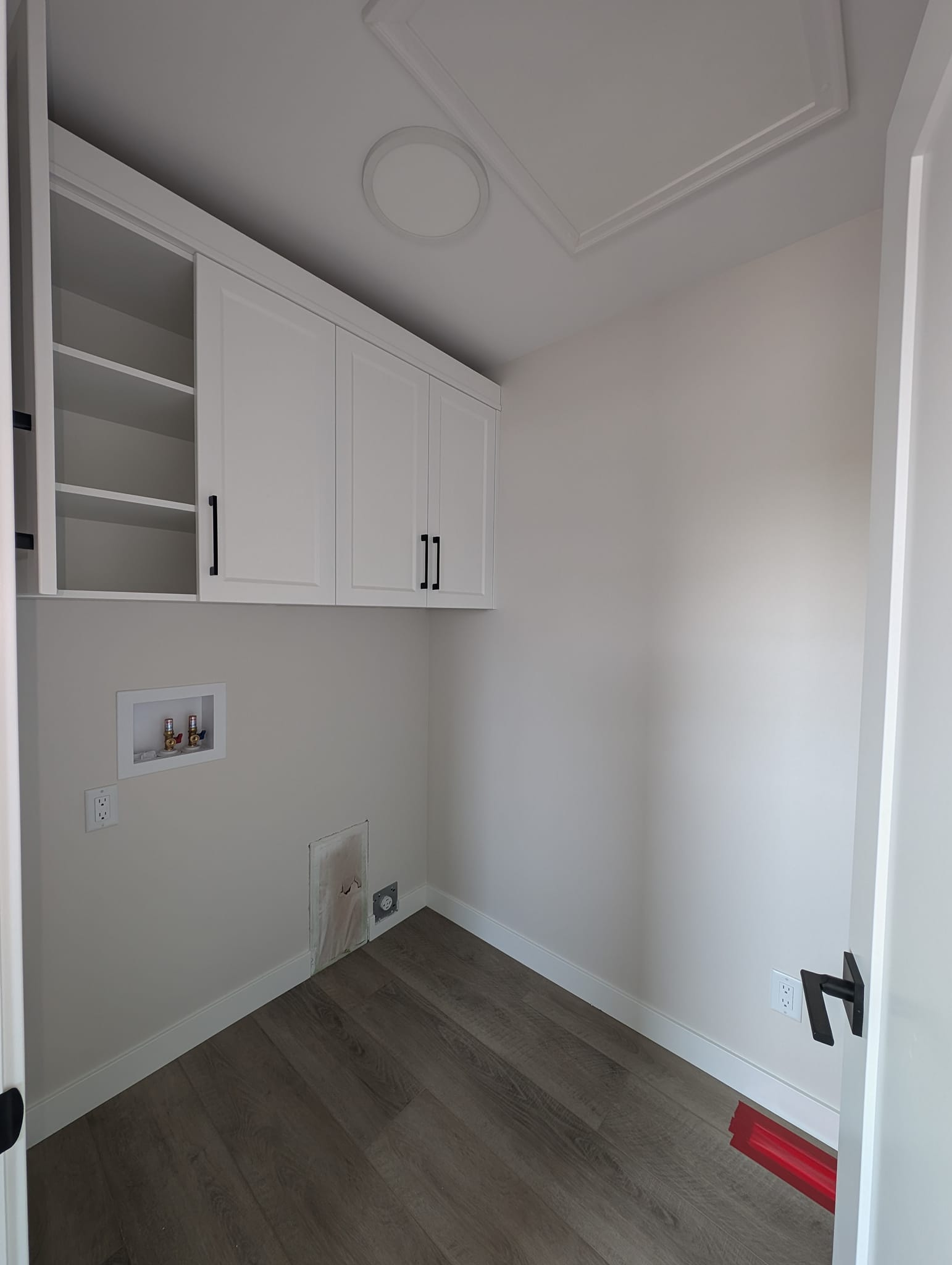 Minimalist laundry room with white walls and wooden floor. Features overhead white cabinets with black handles, plumbing outlets, and ceiling light.