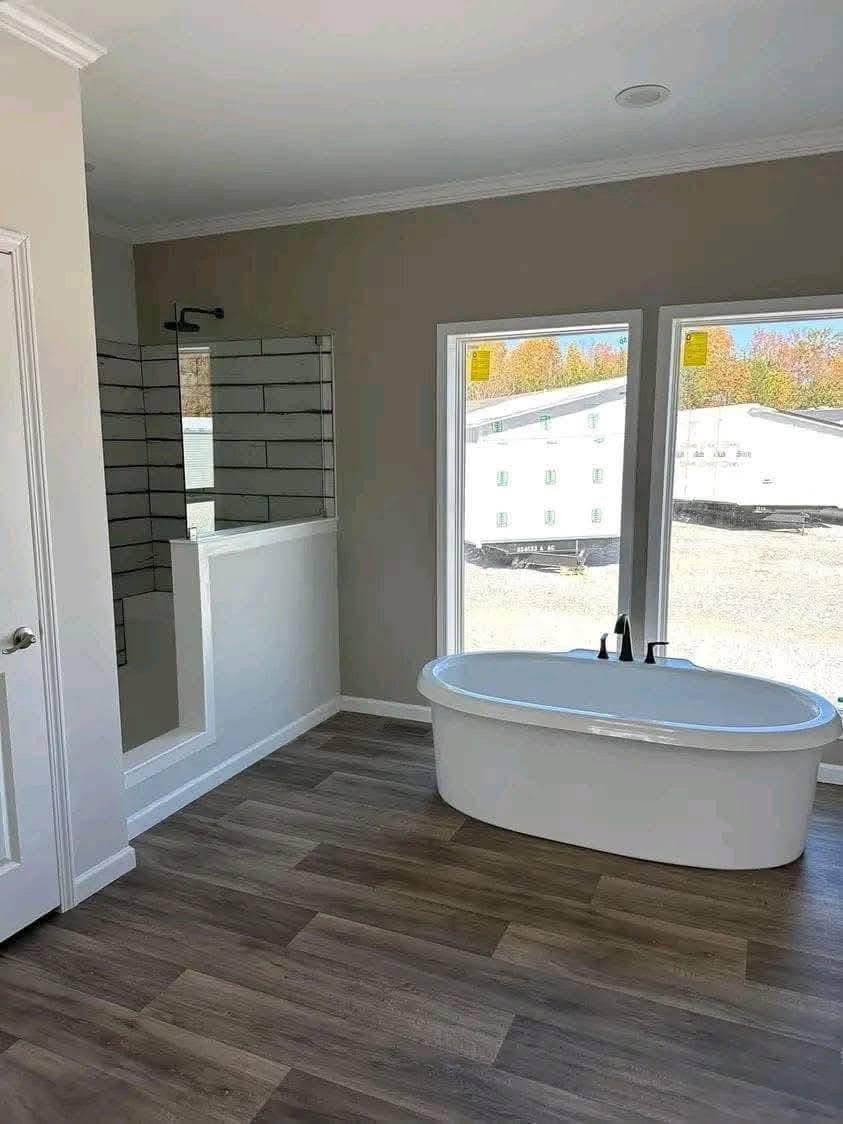 Spacious bathroom with wooden flooring, featuring a sleek white bathtub beside large windows. A modern shower with tiled walls is on the left. Bright and calming ambiance.