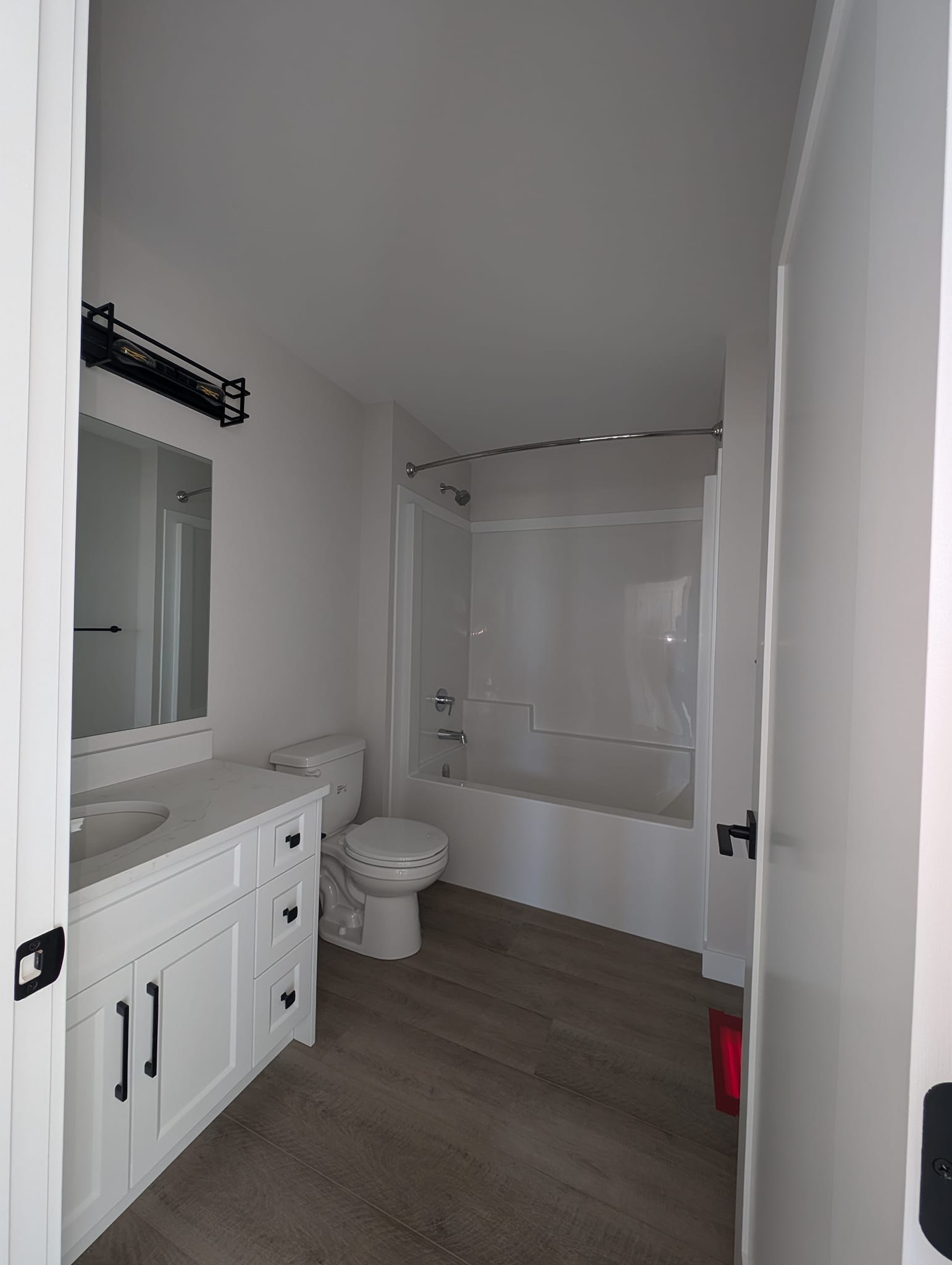 A modern bathroom with white walls and fixtures. It features a bathtub with a curved shower rod, a white vanity with a mirror, and a wooden floor. The space feels clean and minimalistic.