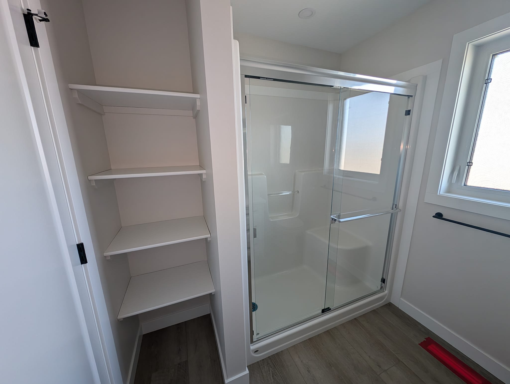 Modern bathroom with a glass-enclosed shower, white shelving unit on the left, and a window on the right. The space is clean and minimalist.