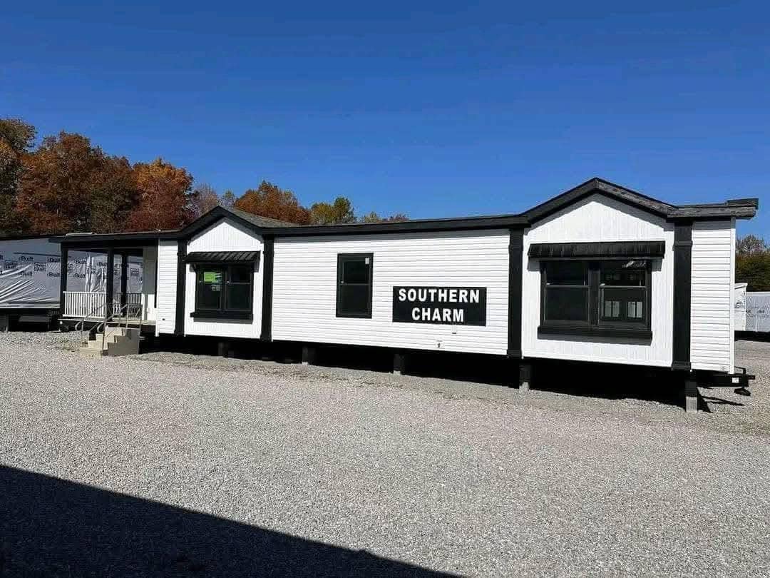 A white modular home labeled "Southern Charm" sits on a gravel lot under a clear blue sky, with autumn trees in the background, creating a cozy feel.