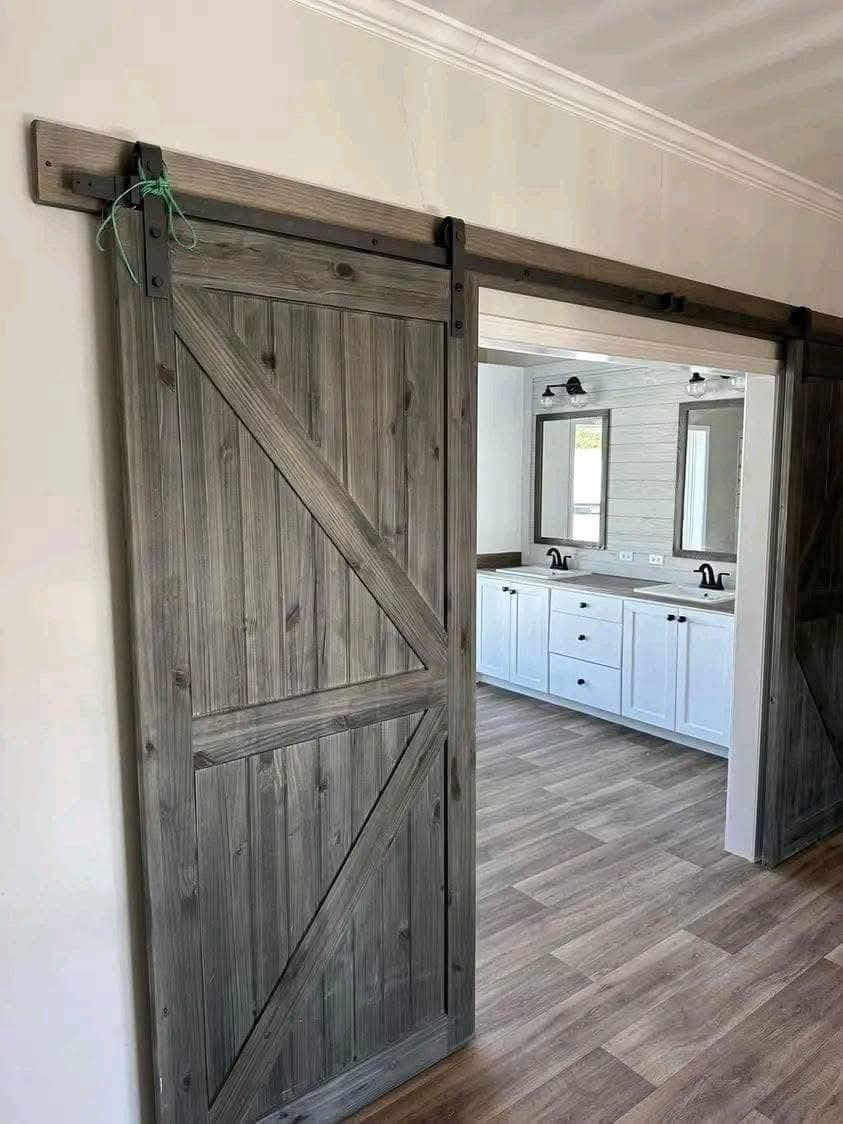 Alt text: "Wooden sliding barn door partially open, revealing a modern bathroom with dual sinks, white cabinets, and mirrors. Rustic and clean aesthetic."