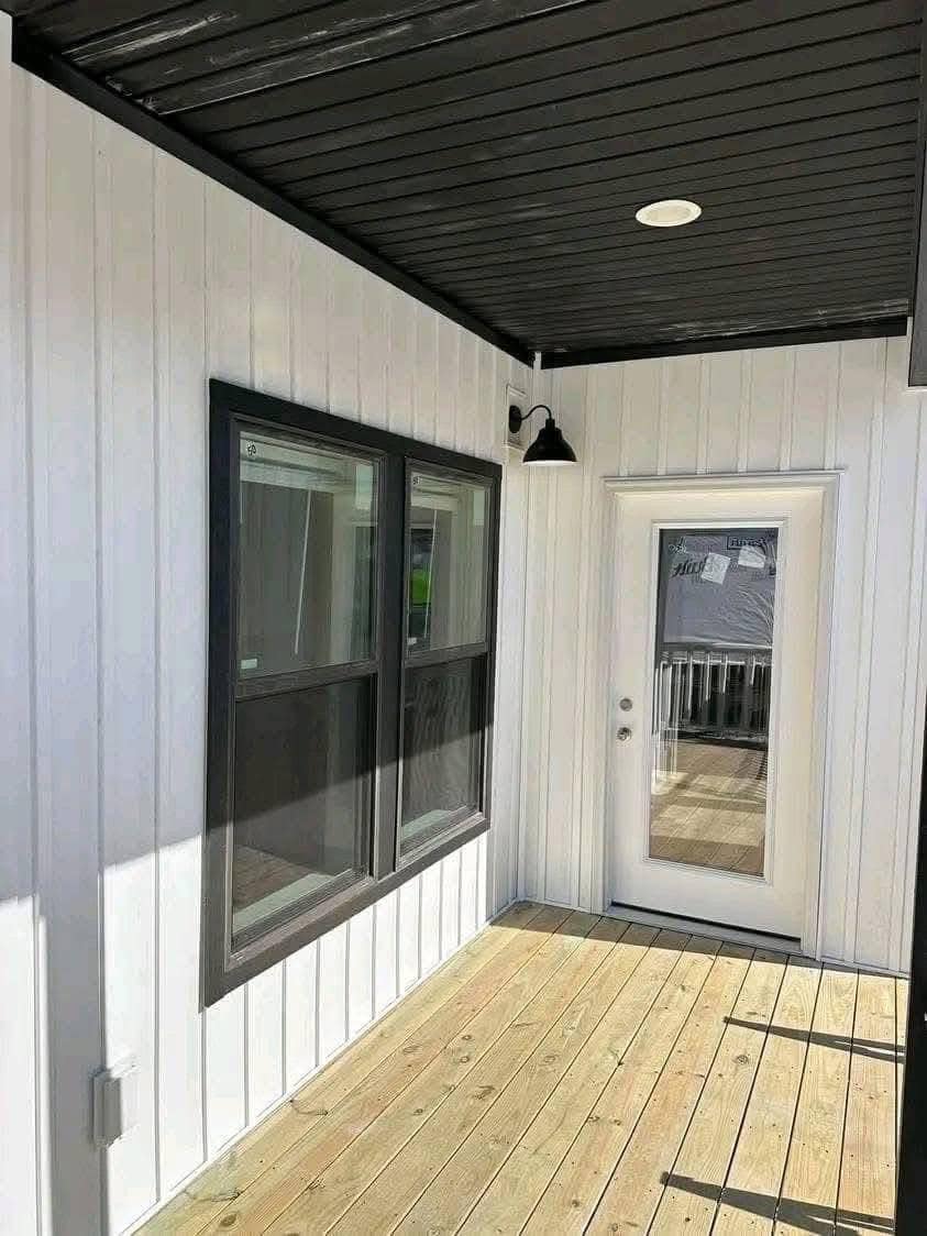 Covered wooden porch with white walls, black accents, and ceiling. Includes a glass door and two windows, creating a modern, clean look.