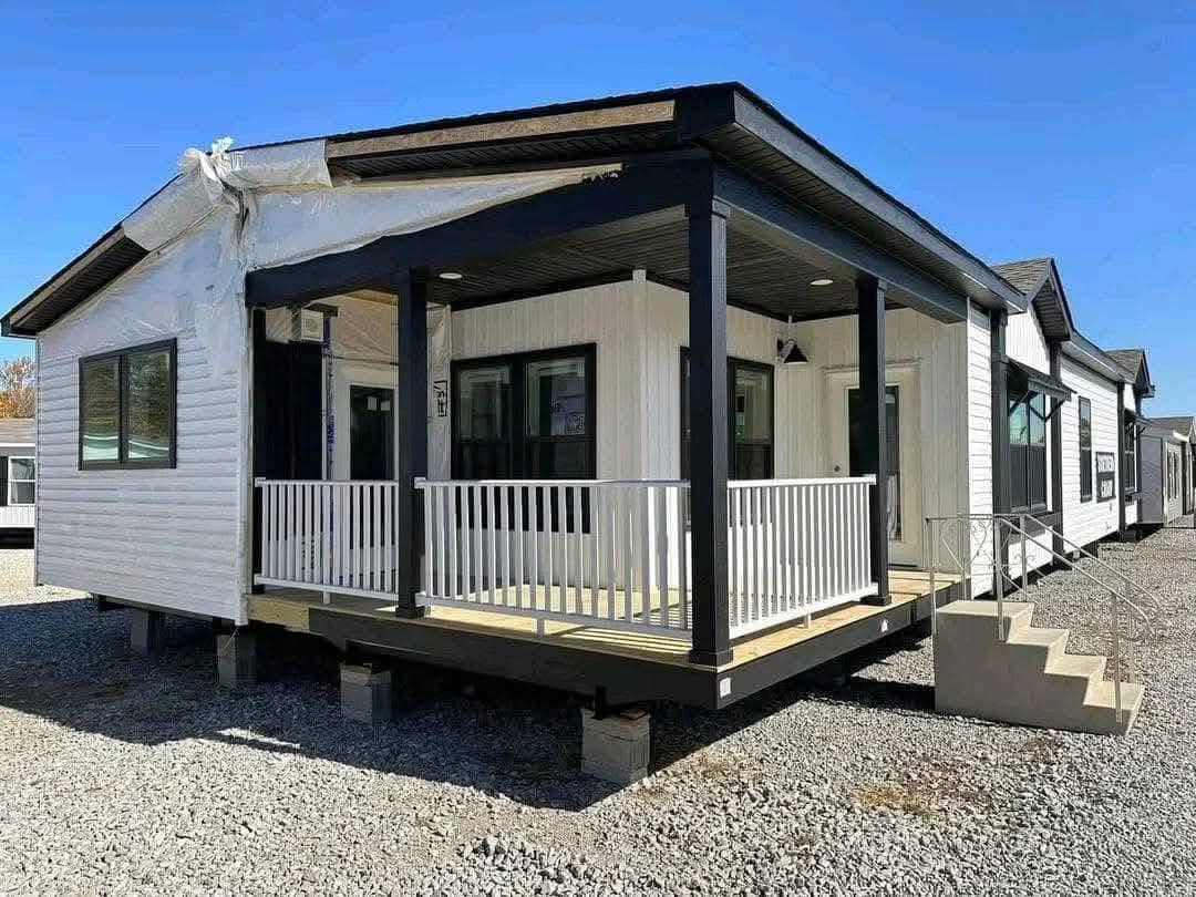 A white modular home with black trim, featuring a covered porch and stairs, sits on gravel under a clear blue sky, conveying a modern, welcoming feel.