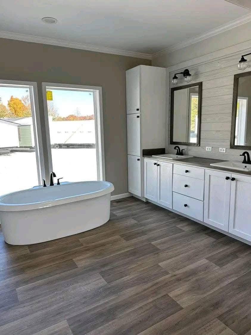 Spacious bathroom with a modern, white freestanding tub, double vanity with two mirrors, black fixtures, wood flooring, and large windows.