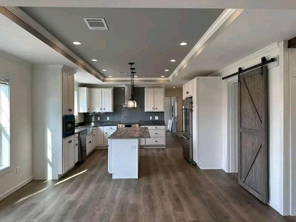 Modern kitchen with wooden flooring, centered island with pendant lights, white cabinets, dark backsplash, and a sliding barn door on the right. Cozy and inviting.