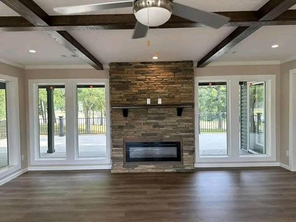 Spacious room with wood flooring and ceiling beams, featuring a stone accent wall with a modern fireplace and large windows offering natural light.