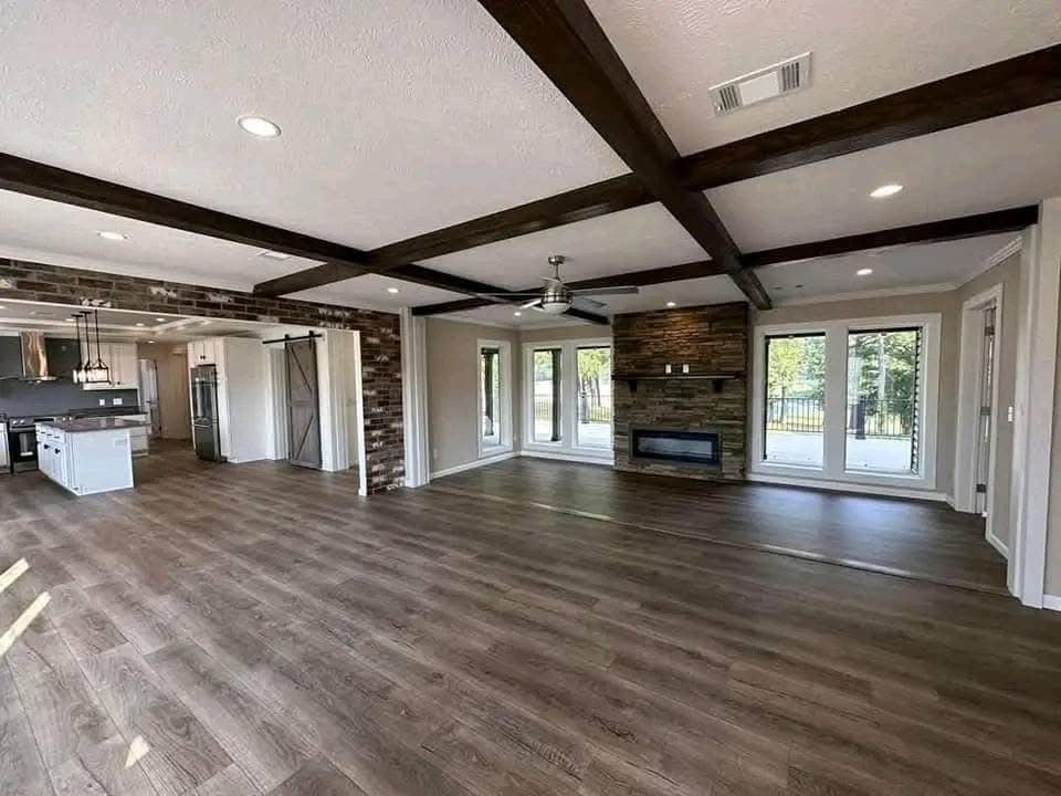 Spacious living room with dark wood beams, light walls, and a stone fireplace. Large windows offer views of greenery, creating an open, airy feel.