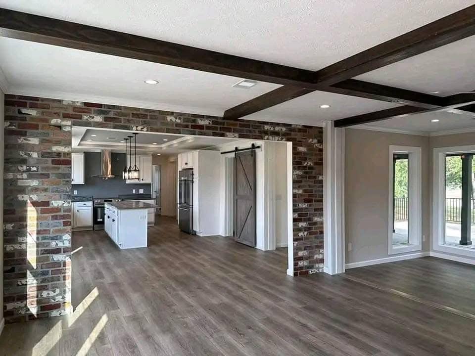 Spacious modern kitchen with wooden floors, exposed beams, and a brick accent wall. Features a large island, barn door, and ample natural light.