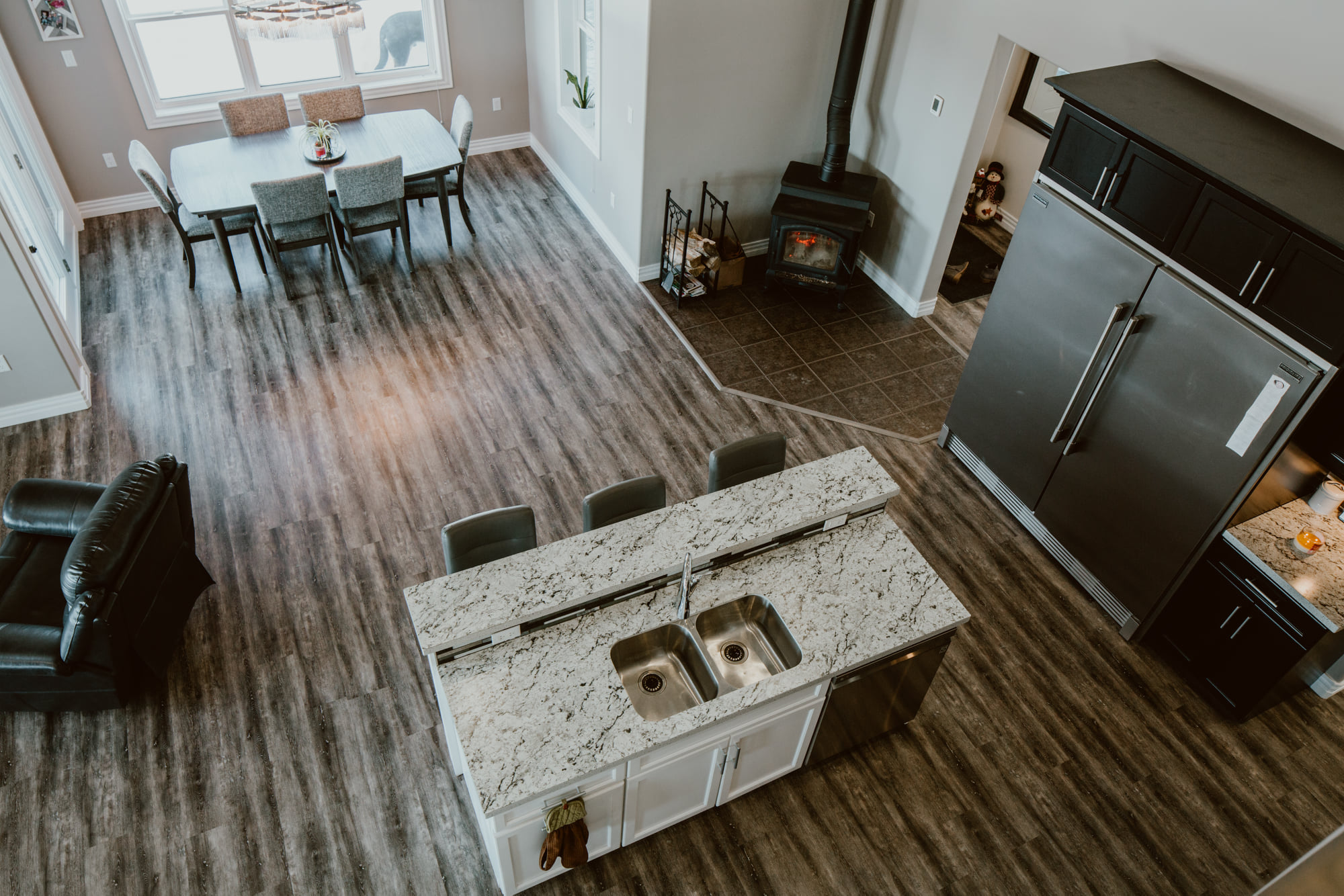 Aerial view of a modern kitchen and dining area. Marble-topped island with sink and stools, wood floors, a dining table by a window, and a cozy wood stove.