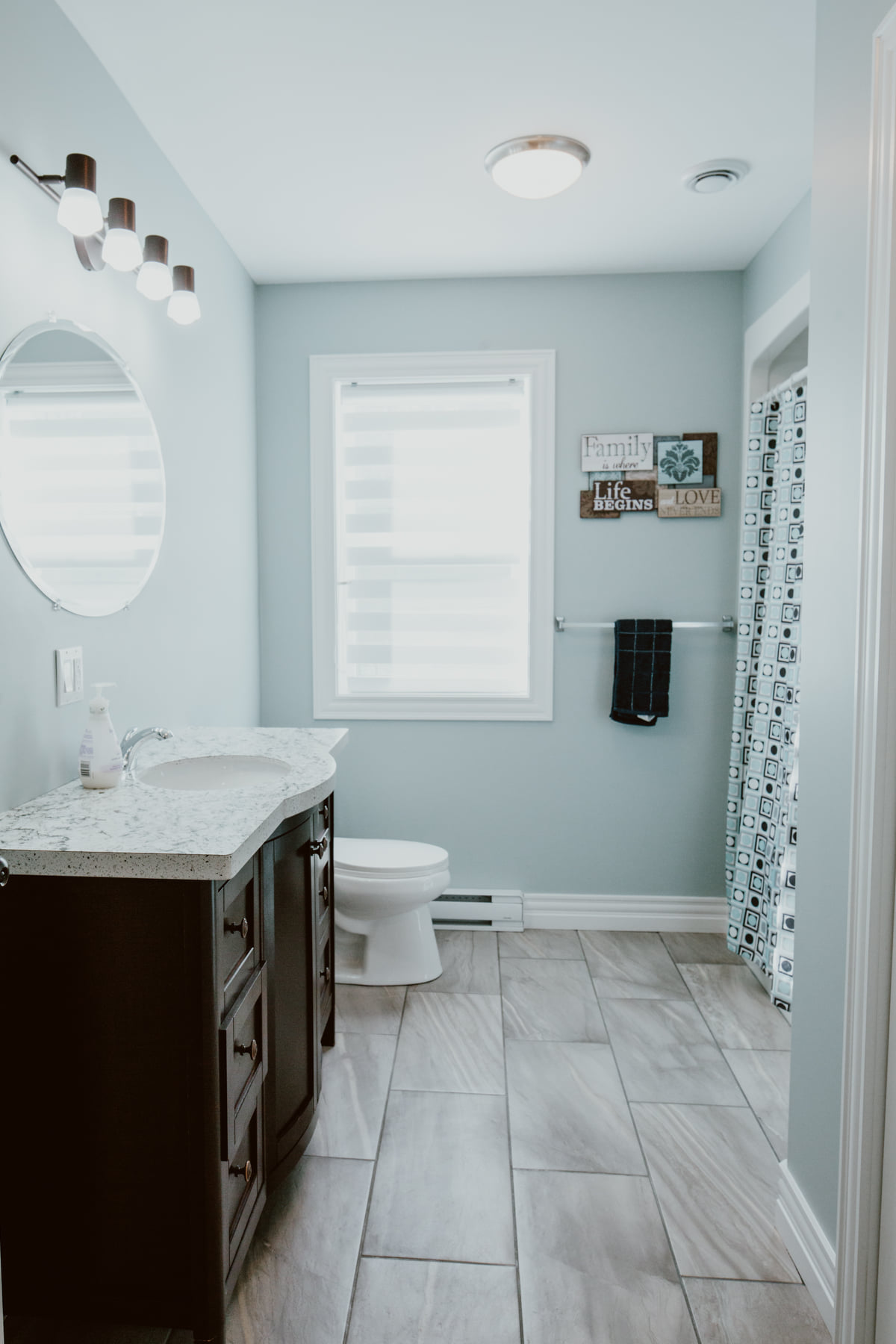 Modern bathroom with light blue walls and tiled floor. Features a dark wood vanity with a marble countertop, round mirror, and bright overhead lights. A window with sheer blinds is above the toilet, next to wall art reading "family" and "love". A patterned shower curtain is visible on the right. Cozy and clean atmosphere.
