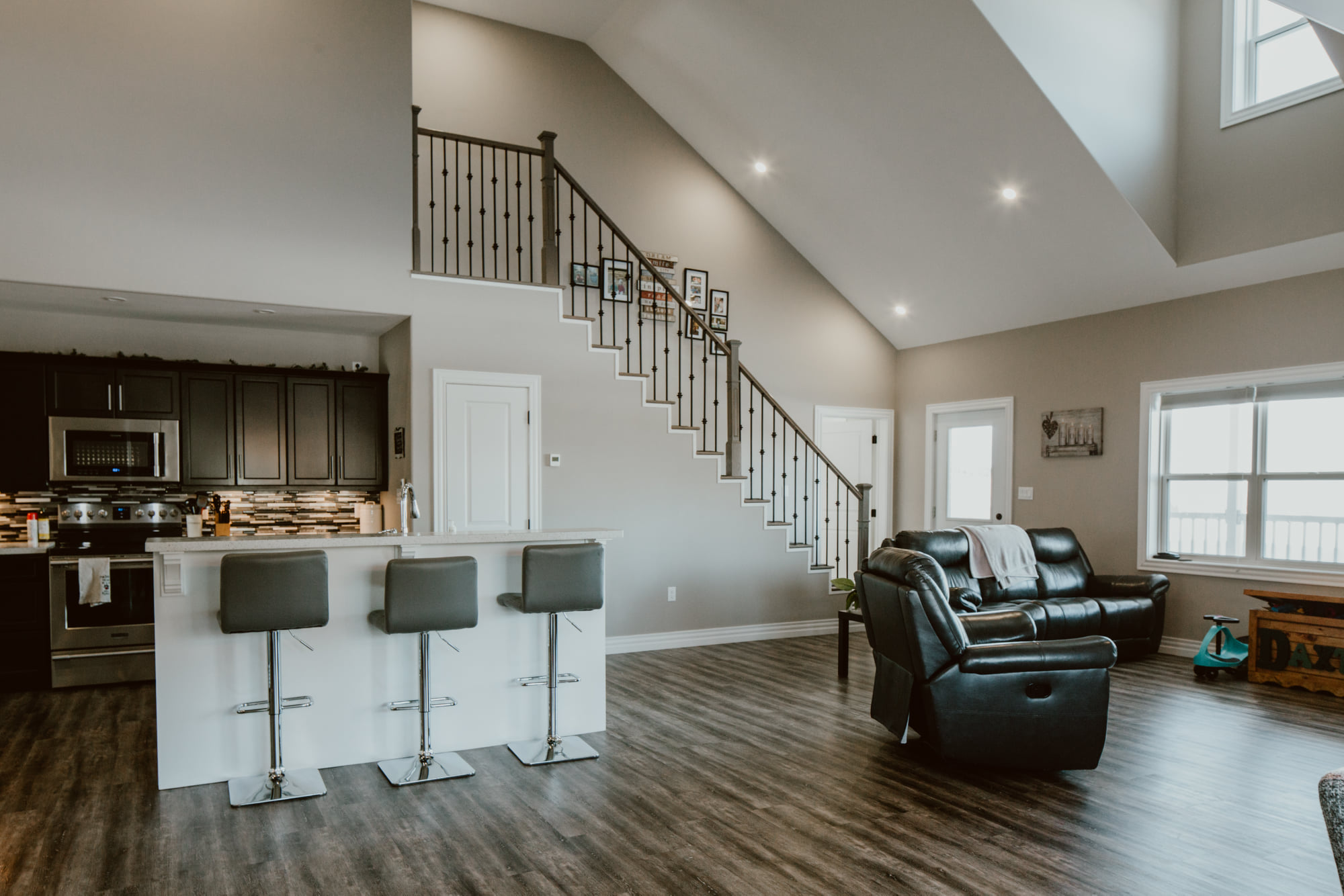 Modern living room with dark wood floors, black cabinets, and a kitchen island with bar stools. A staircase with iron railings accentuates the space.