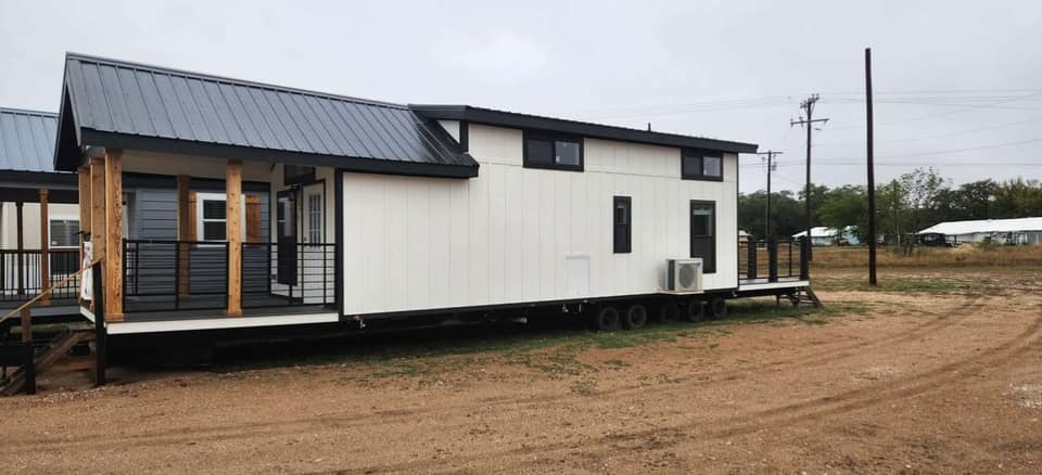 A modern tiny house on wheels with a black metal roof and white siding sits on a dirt lot. It features wooden accents and a small porch, conveying a minimalist and cozy feel.
